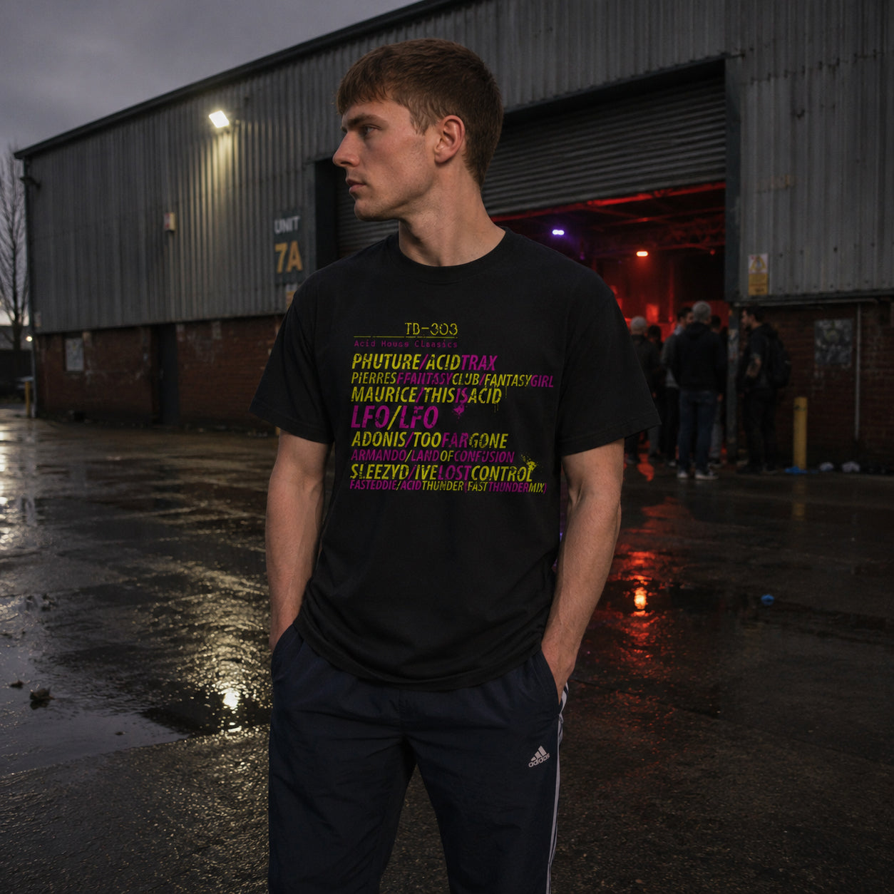 A man wearing a black graphic t-shirt and dark athletic pants stands in a wet outdoor industrial yard in front of a large metal warehouse door. The t-shirt features Acid House Roland synth graphic tee