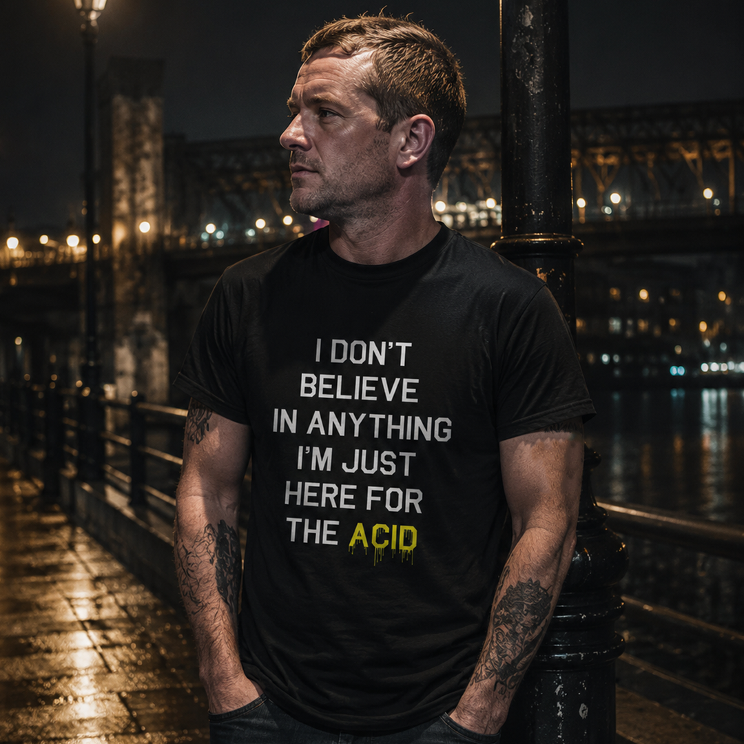 A man wearing a black graphic t-shirt with the text 'I DON'T BELIEVE IN ANYTHING I'M JUST HERE FOR THE ACID' in white and yellow, standing on a wet city bridge at night.
