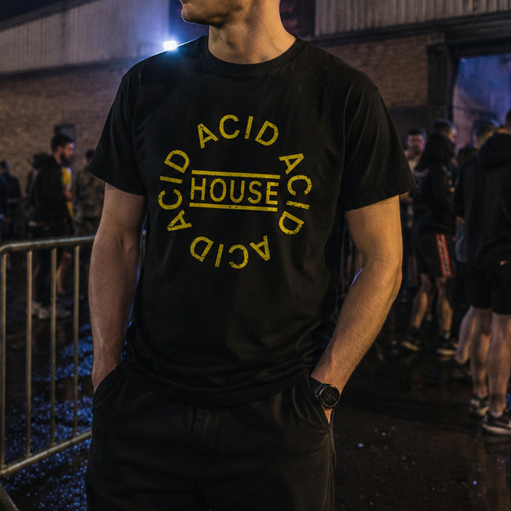 A man wearing a black t-shirt with a yellow 'ACID HOUSE' graphic print, standing in a dimly lit outdoor or event setting at night.