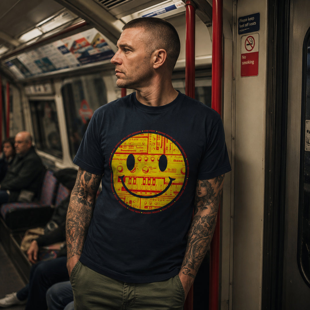 A man with tattoos stands on a subway train wearing a navy blue t-shirt with a large yellow and red retro tb-303 smiley face graphic on the front.