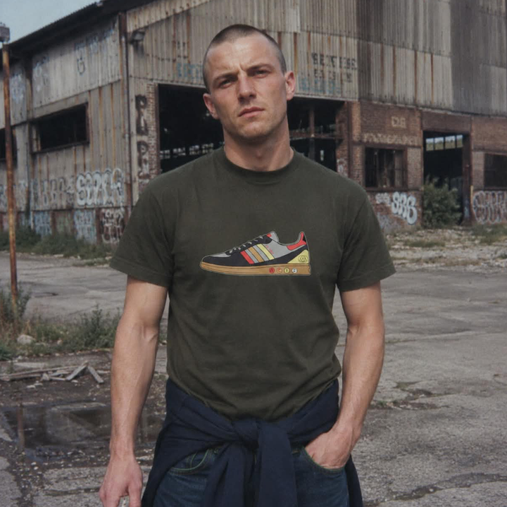 A man wearing an olive green t-shirt with a graphic of a black and red Adidas Samba sneaker, standing in front of a weathered, graffiti-covered industrial building.