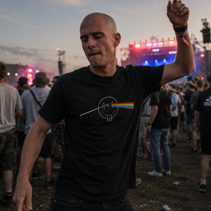 A man at a concert wearing a black t-shirt with a white circular graphic and a rainbow light beam from the Dark side of the Moon, standing in a crowd with stage lights in the background.