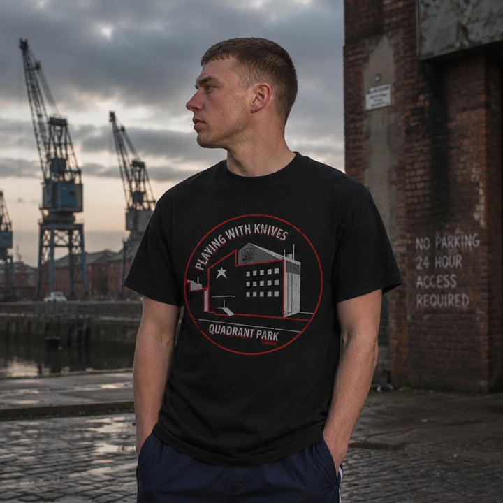 A man wearing a black graphic t-shirt with a red and white 'Playing with Knives Quadrant Park' design, standing in an outdoor harbor setting with cranes and a brick wall in the background.