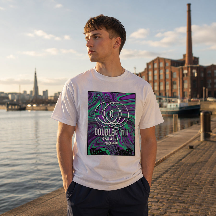 A young man standing on a waterfront promenade wearing a white crewneck t-shirt with a colorful psychedelic graphic print on the front. The graphic features a purple and green psychedelic swirl pattern with the text 'Double Good Garments'.
