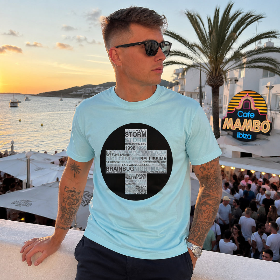 A man wearing a light blue Trance Classics t-shirt with a black circular graphic containing text, standing outdoors on a balcony overlooking a beach and Ibiza sunset with a crowded Cafe Mambo in the background.